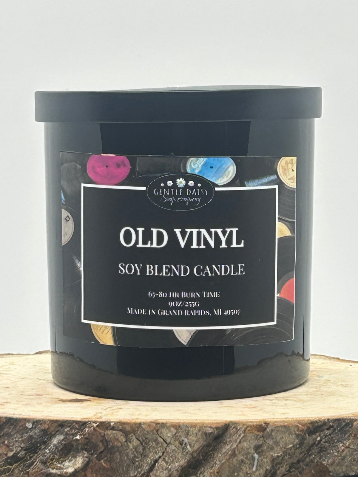 A black candle jar labeled 'OLD VINYL SOY BLEND CANDLE' by Ghost Daisy Candle Company, placed on a rustic wood slice against a light gray background.