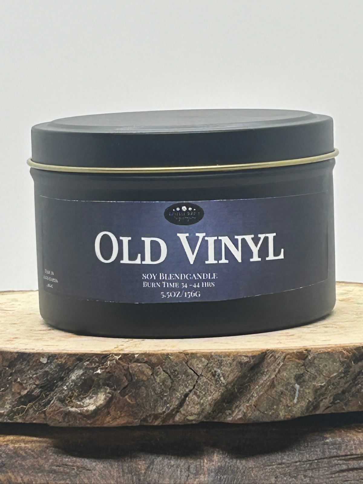 A dark round metal tin candle labeled 'OLD VINYL' sits on a rustic wood slice against a plain light background.