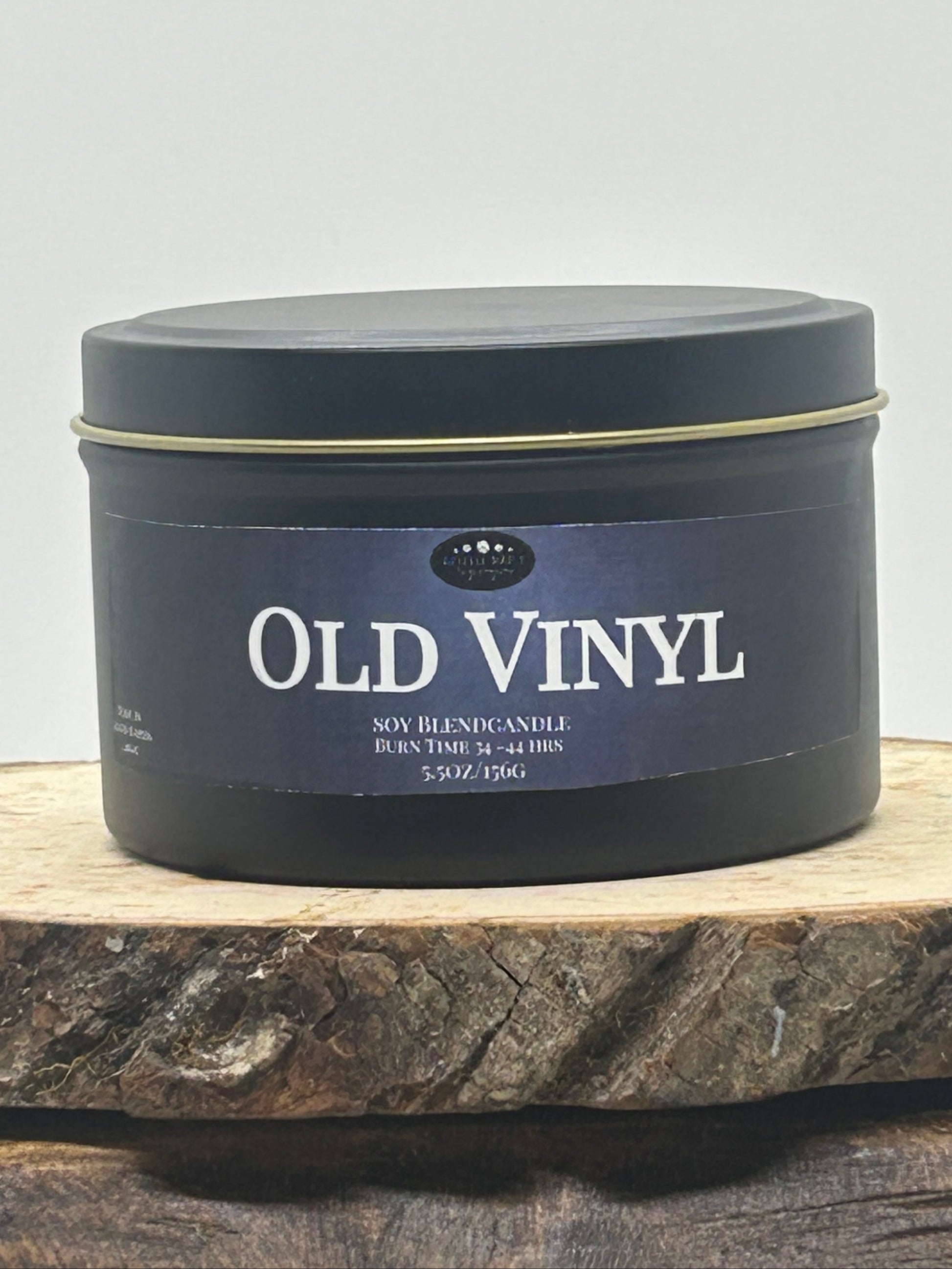 A dark round metal tin candle labeled 'OLD VINYL' sits on a rustic wood slice against a plain light background.