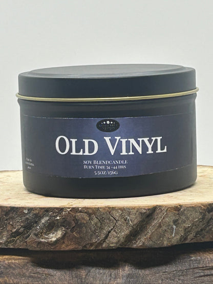 A dark round metal tin candle labeled 'OLD VINYL' sits on a rustic wood slice against a plain light background.