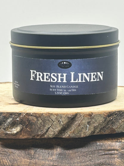 A dark navy blue metal tin candle labeled 'FRESH LINEN' sits on a rustic wood slice against a light gray background.