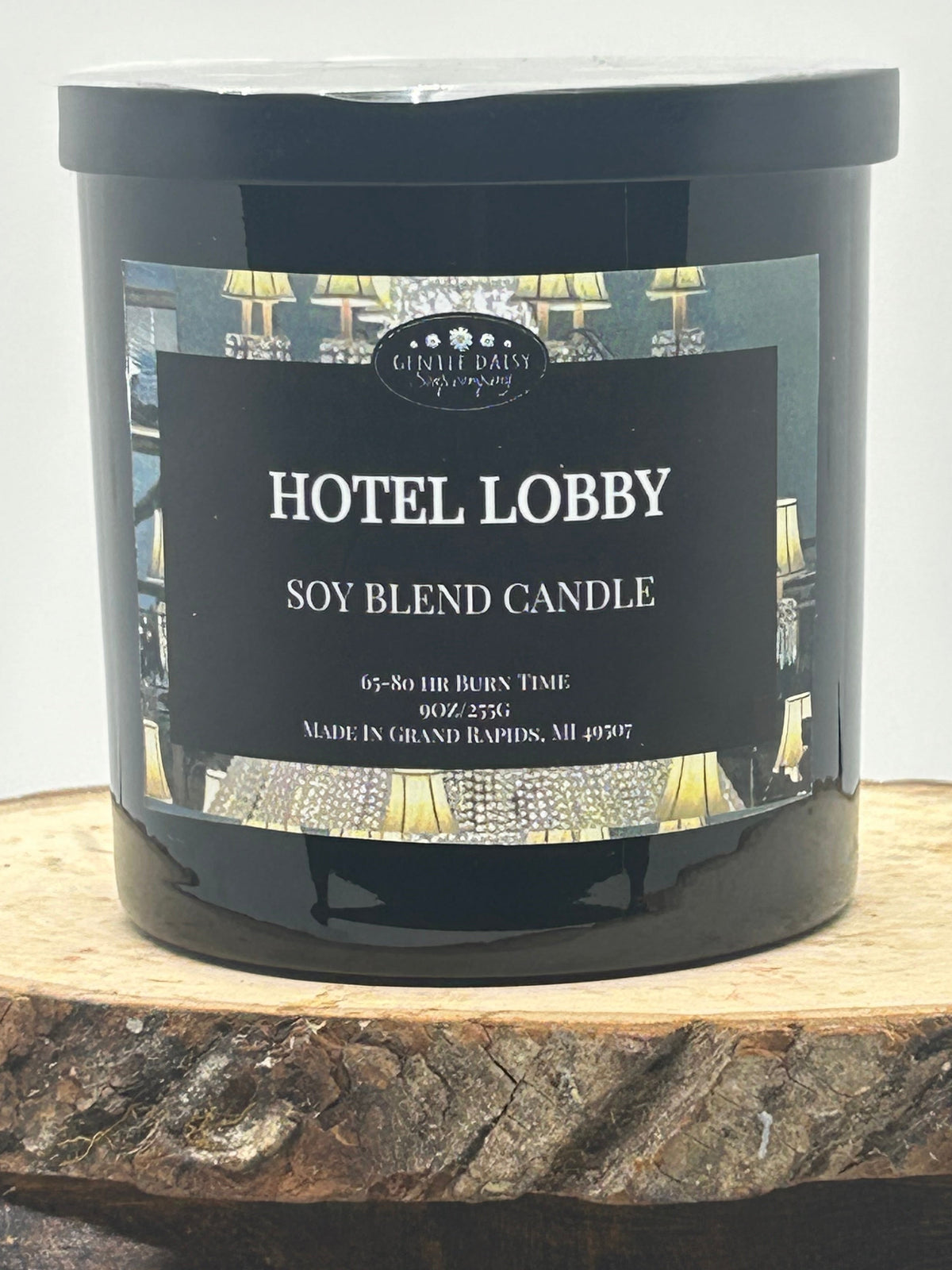 A black candle jar labeled 'Hotel Lobby Soy Blend Candle' by Candle Creek Lighting, displayed on a rustic wood slice against a light gray background.