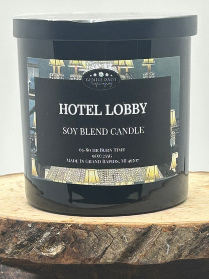 A black candle jar labeled 'Hotel Lobby Soy Blend Candle' by Candle Creek Lighting, displayed on a rustic wood slice against a light gray background.
