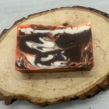 A rectangular bar of marbled soap with dark brown, black, and white swirls and orange-red edges, displayed on a round natural wood slice against a light gray textured background.