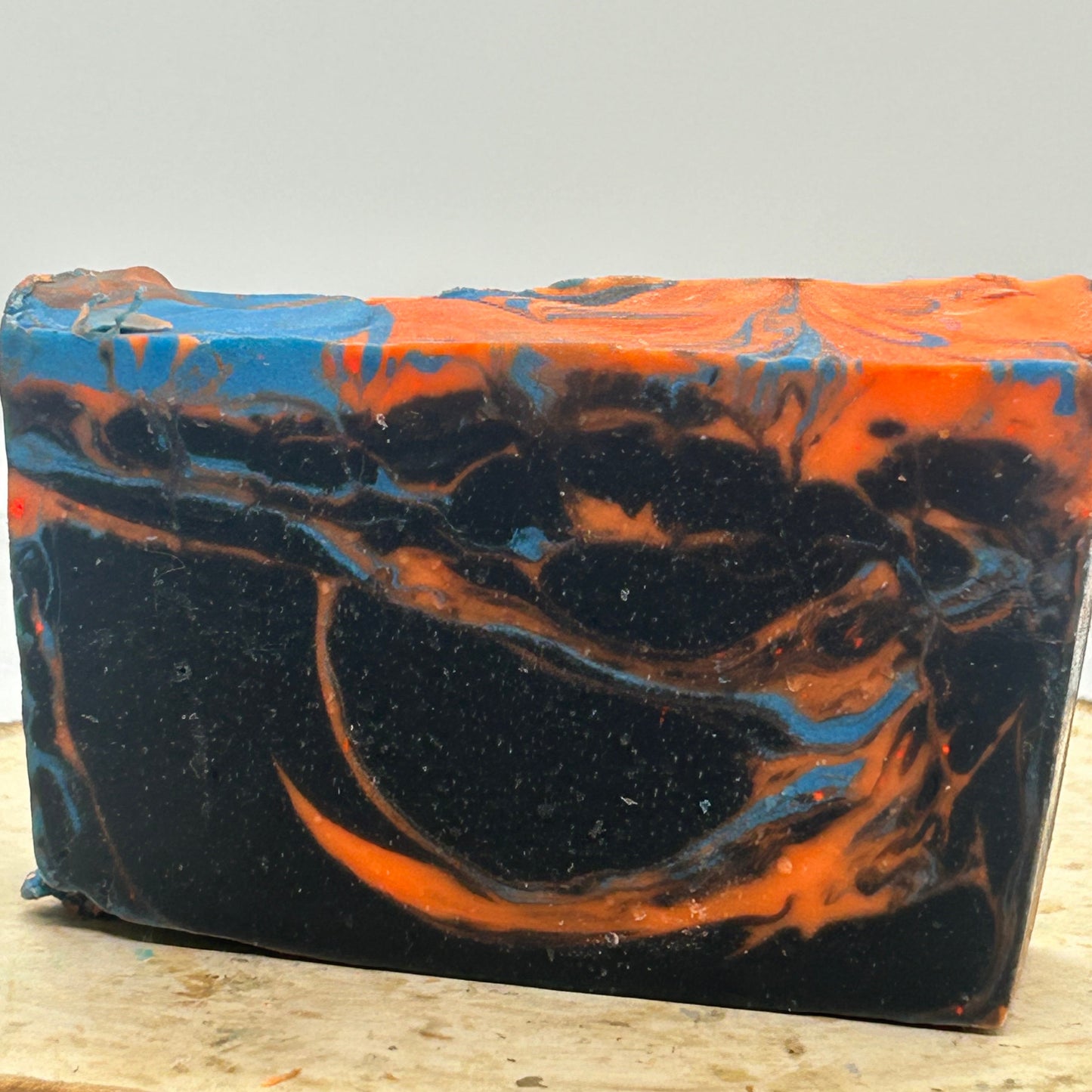 A rectangular handmade soap bar with a marbled swirl pattern of black, bright orange, and blue, displayed on a rustic wood slice against a neutral light gray background.