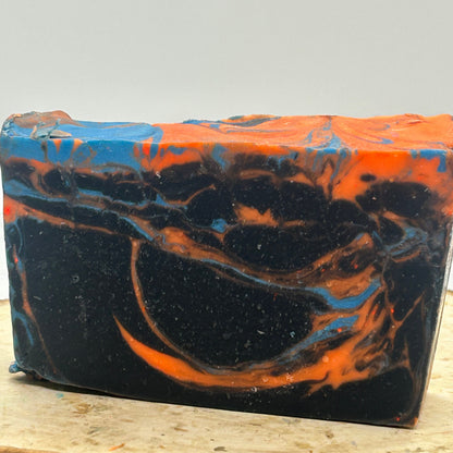 A rectangular handmade soap bar with a marbled swirl pattern of black, bright orange, and blue, displayed on a rustic wood slice against a neutral light gray background.
