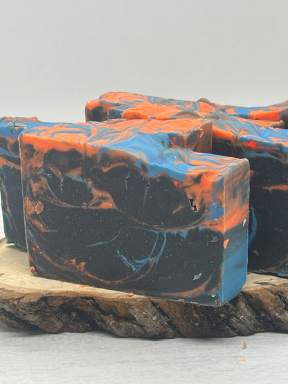 Three rectangular bars of marbled soap with a black base, bright blue swirls, and orange accents, displayed on a rustic wood slice against a light gray background.