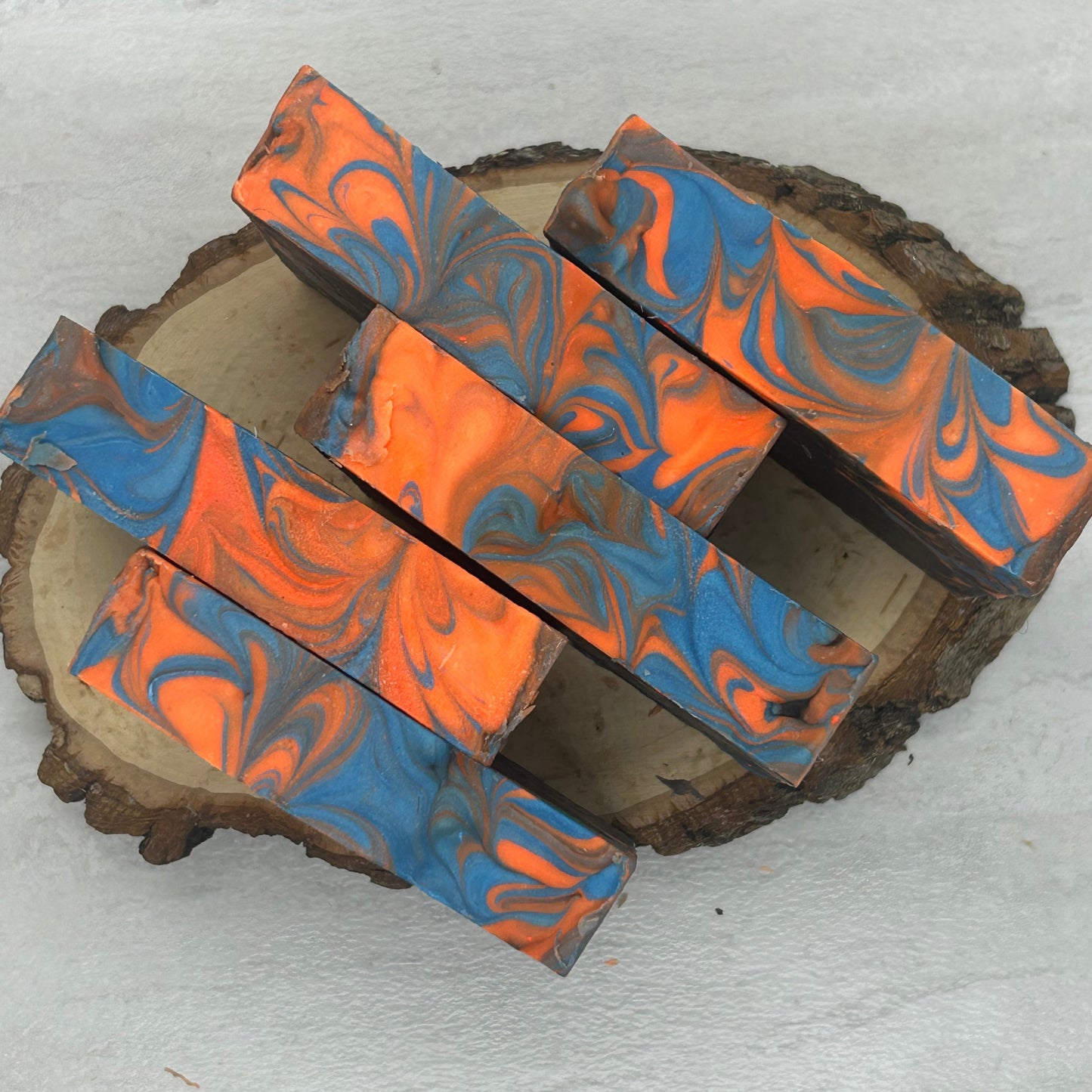 Five rectangular bars with a blue and orange marbled swirl pattern, arranged on a rustic round wood slice against a light gray textured background.